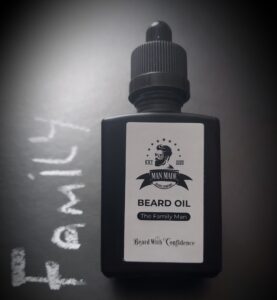 A short rectangular glass Bottle with a black rubber pipette lid and a light brown label that has Man Made Beard Company The Family Man Beard Oil written in bold black writing and a picture of a man with a black beard on it, on a white background.
