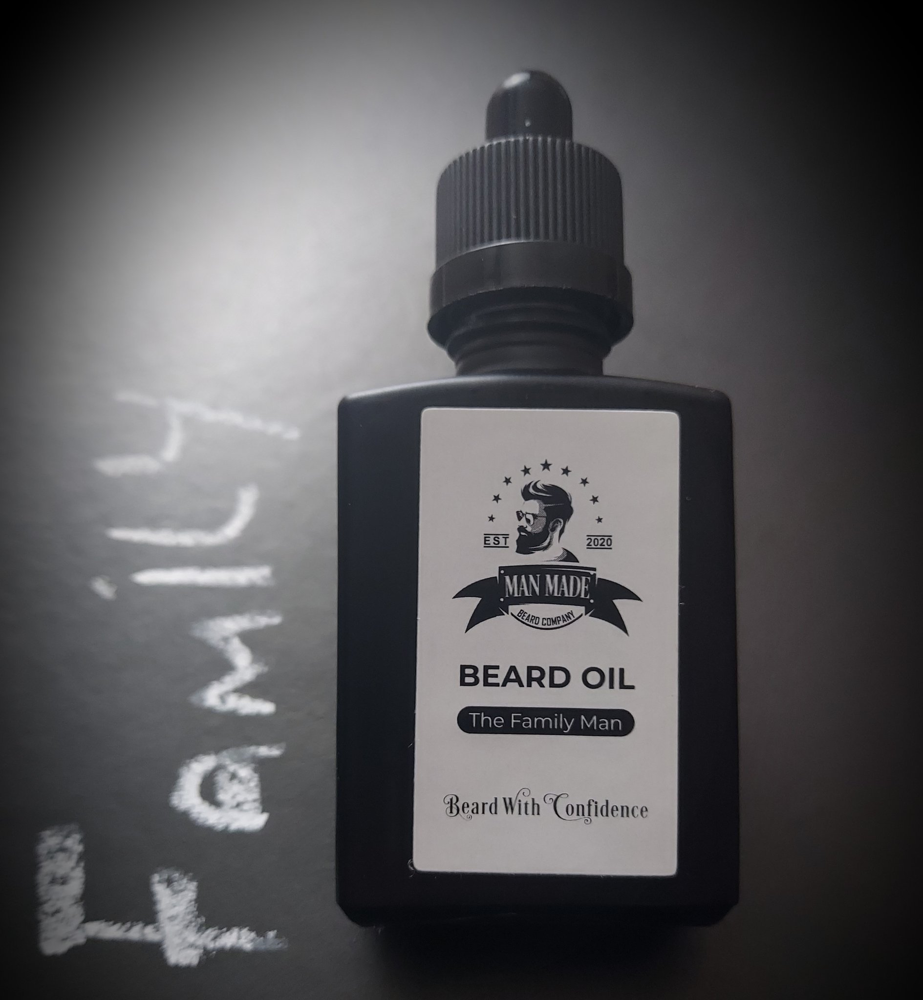 Lucky There's A Family Man | Man Made Beard Company The Family Man Beard Oil A short rectangular glass Bottle with a black rubber pipette lid and a light brown label that has Man Made Beard Company The Family Man Beard Oil written in bold black writing and a picture of a man with a black beard on it, on a white background.