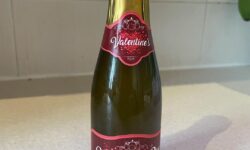 A short cylindrical clear glass wine bottle full of some fizzy liquid with a gold foil lid and a pale purple label that has Valentines Day written in bold white writing and Luke Sam Sowden, Bubbles, Bubbles, Bubbles, He likes bubbles written in bold gold writing on it, on a white background.