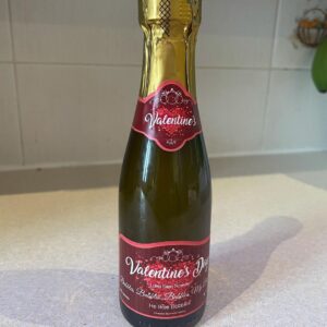 A short cylindrical clear glass wine bottle full of some fizzy liquid with a gold foil lid and a pale purple label that has Valentines Day written in bold white writing and Luke Sam Sowden, Bubbles, Bubbles, Bubbles, He likes bubbles written in bold gold writing on it, on a white background.