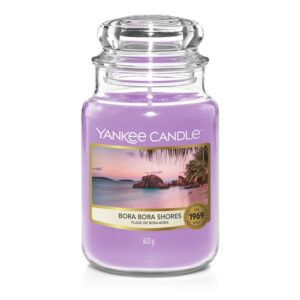 A tall cylindrical Jar full of some sandy yellow Wax with a label that has Yankee Candle written in white writing, bora bora shores written in black writing, and a picture of a white rocky cliff overlooking a sandy beach on it, on a white background.