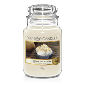 A tall cylindrical Jar full of some creamy white Wax with a label that has Yankee Candle written in white writing, coconut rice dream written in black writing, and a picture of a dark brown half a coconut full of some creamy white cream on it, on a white background.