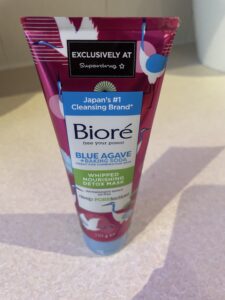 A tall conical bright pink plastic tube with a bright blue lid and a bright white label that has Biore Blue Agave & Baking Soda Whipped Detox Mask written in dark blue writing and some pictures of some storks on it, on a white background.