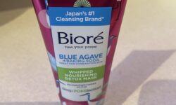 A tall conical bright pink plastic tube with a bright blue lid and a bright white label that has Biore Blue Agave & Baking Soda Whipped Detox Mask written in dark blue writing and some pictures of some storks on it, on a white background.