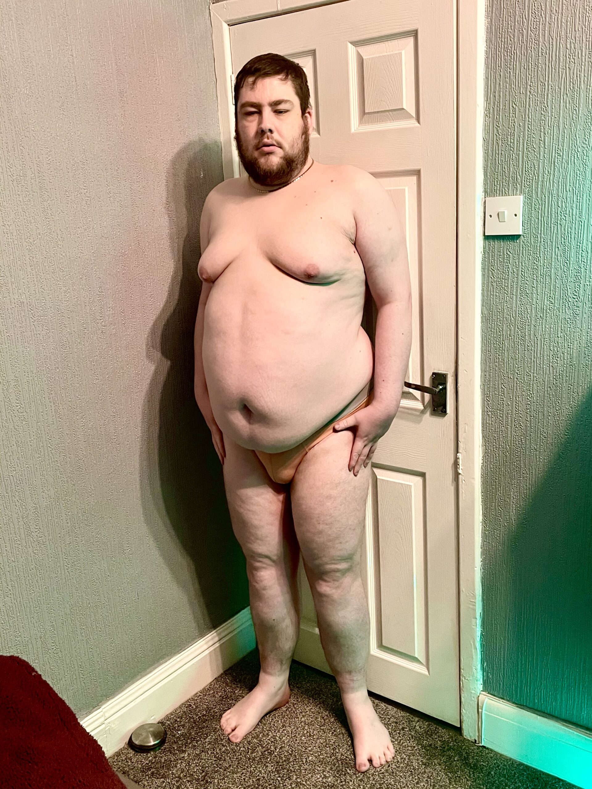 A tall bearded man wearing a pair of peach briefs with a peach waistband that has Blanc Menswear written in bold black writing on it stood against a white wooden wall.
