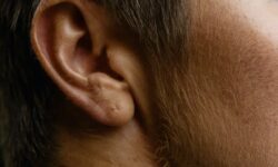 A tall white man wearing a navy blue shirt that has his Head to the side so that you can see his ear, on a white background.