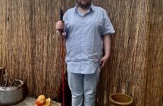 A tall bearded man wearing a light blue short sleeve shirt that has a white diamond print all over it, a pair of sky blue chino trousers and a pair of shiny red shoes holding a bright red cane stood against a dark wooden fence.