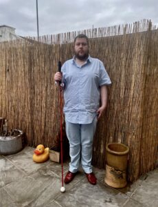 A tall bearded man wearing a light blue short sleeve shirt that has a white diamond print all over it, a pair of sky blue chino trousers and a pair of shiny red shoes holding a bright red cane stood against a dark wooden fence.