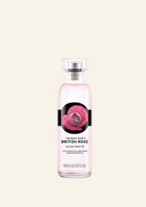 A tall cylindrical clear glass bottle full of some translucent pink liquid with a metallic silver lid and a black label that has The Body Shop British Rose Eau De Toilette written in bold black writing and some pictures of some bright pink rose petals on it, on a white background.