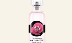A tall cylindrical clear glass bottle full of some translucent pink liquid with a metallic silver lid and a black label that has The Body Shop British Rose Eau De Toilette written in bold black writing and some pictures of some bright pink rose petals on it, on a white background.