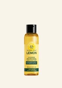 A short cylindrical clear plastic bottle with a black plastic lid and a bright yellow label that has The Body Shop Lemon Cleansing Anti-Bacterial Hand Sanitiser written in dark green writing, a dark green stripe, and a picture of a bright yellow lemon on it, on a white background.