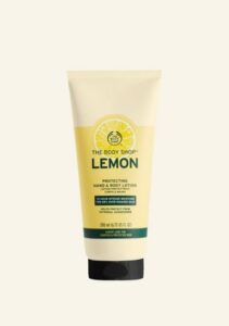 A tall conical white plastic tube with a black metal lid and a bright yellow label that has The Body Shop Lemon Protecting Hand & Body Lotion written in dark green writing, a dark green stripe, and a picture of a bright yellow lemon on it, on a white background.