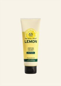 A tall conical white plastic tube with a black metallic lid and a bright yellow label that has The Body Shop Lemon Purifying Face Wash written in dark green writing, a dark green stripe, and a picture of a bright yellow lemon on it, on a white background.