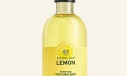 A tall cylindrical clear plastic bottle full of some bright yellow liquid with a bright yellow label that has The Body Shop Lemon Purifying Hair & Body Wash written in dark green writing, a dark green stripe, and a picture of a bright yellow lemon on it, on a white background.