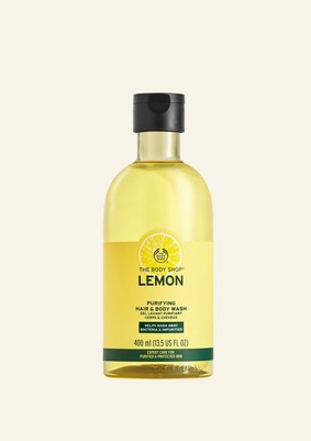 A tall cylindrical clear plastic bottle full of some bright yellow liquid with a bright yellow label that has The Body Shop Lemon Purifying Hair & Body Wash written in dark green writing, a dark green stripe, and a picture of a bright yellow lemon on it, on a white background.