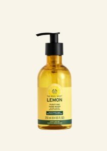 A tall rectangular clear plastic bottle full of some bright yellow liquid with a black pump lid and a bright yellow label that has The Body Shop Lemon Purifying Hand Wash written in dark green writing, a dark green stripe, and a picture of a bright yellow lemon on it, on a white background.
