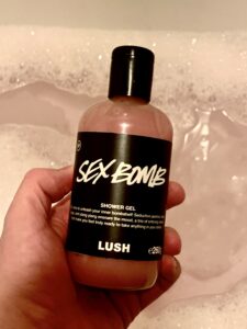 A hand holding a tall cylindrical clear plastic bottle full of some light pink liquid with a black label that has Lush Sex Bomb Shower Gel written in bold white writing on it, on a white background.