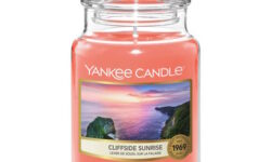 A tall cylindrical Glass Jar full of some sandy yellow wax with a label that has Yankee Candle written in white writing, Cliffside Sunrise written in black writing, and a picture of a white rocky cliff surrounded by the sea on it, on a white background.