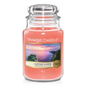 A tall cylindrical Glass Jar full of some sandy yellow wax with a label that has Yankee Candle written in white writing, Cliffside Sunrise written in black writing, and a picture of a white rocky cliff surrounded by the sea on it, on a white background.