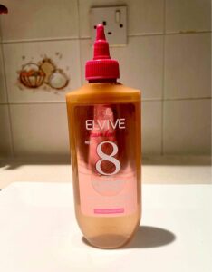 A tall rounded rectangular clear plastic bottle full of some clear liquid with a pink plastic nozzle lid that has L’Oreal Paris Elvive Dream Lengths Wonder Water written in bold white writing on it, on a white background.
