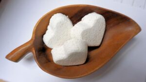 A small bright white heart shaped bath bomb that has some pieces of epsom salt embedded in it, on a white background.