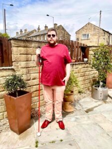 A tall bearded man wearing a pair of silver metal Aviator Sunglasses with some light grey lenses, a bright red crew neck t-shirt, a pair of pale pink chino trousers and a pair of shiny red shoes holding a bright red cane stood against a dark wooden fence.
