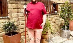 A tall bearded man wearing a pair of silver metal Aviator Sunglasses with some light grey lenses, a bright red crew neck t-shirt, a pair of pale pink chino trousers and a pair of shiny red shoes holding a bright red cane stood against a dark wooden fence.