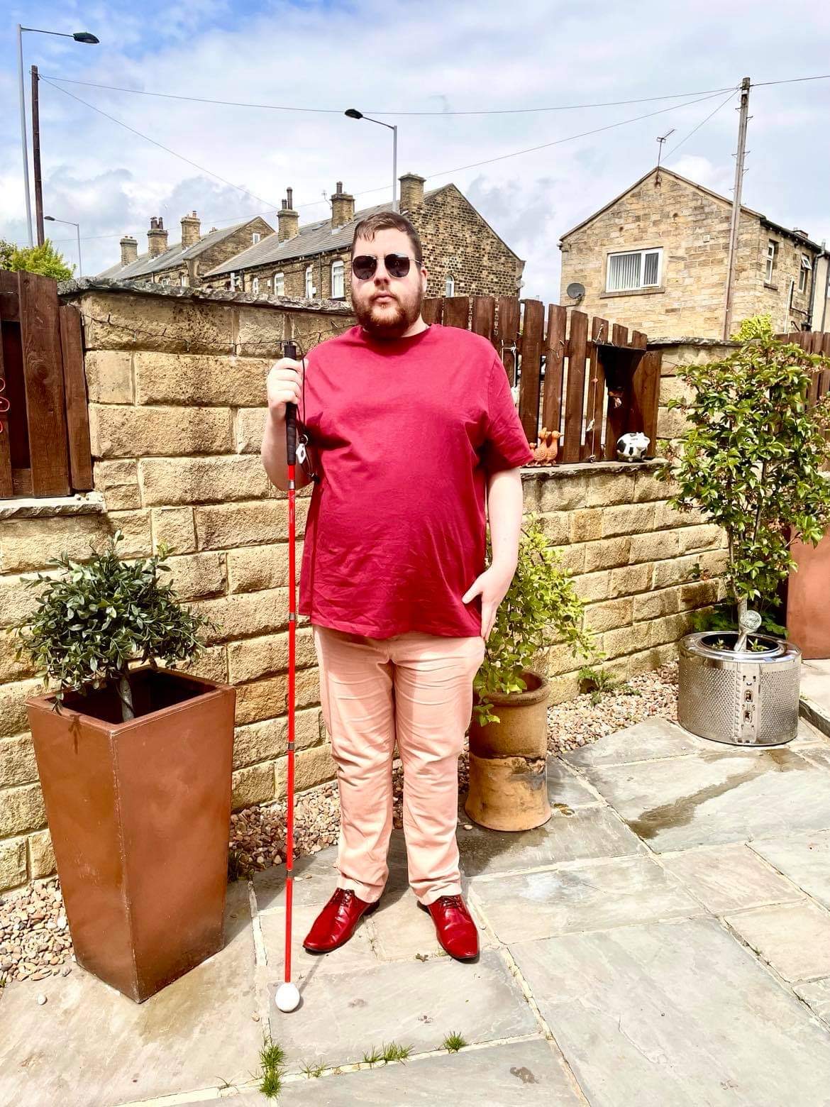 Outfit Of The Day | Receiving My Second COVID-19 Vaccine A tall bearded man wearing a pair of silver metal Aviator Sunglasses with some light grey lenses, a bright red crew neck t-shirt, a pair of pale pink chino trousers and a pair of shiny red shoes holding a bright red cane stood against a dark wooden fence.