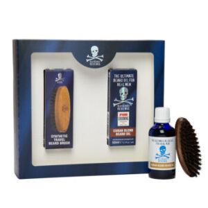 A large rectangular bright blue and white cardboard box that has The Bluebeards Revenge Cuban Beard Care Grooming Kit written in bold white writing on it next to a short cylindrical brown glass bottle and a large oval dark wooden beard brush, on a white background.