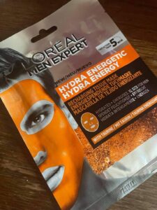 A tall rectangular silver plastic pouch that has L’Oreal Paris Men Expert Hydra Energetic Tissue Mask written in bold black writing and a picture of a mans face that's wearing a bright orange face mask on it, on a white background.