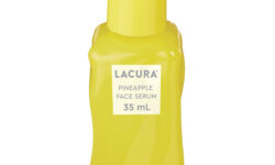 A tall rectangular yellow glass bottle full of some clear liquid with a yellow rubber pipette lid and a white label that has Aldi Lacura Pineapple Face Serum written in bold black writing on it, on a white background.