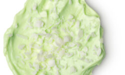 A medium circular puddle of light green body butter that has some chunks of shea butter running through it, on a white background.