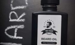 A short rectangular brown glass Bottle with a black rubber pipette lid and a white label that has Man Made Beard Company The Hard Man Beard Oil written in bold black writing and a picture of a man with a black beard on it, on a white background.