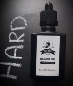 A short rectangular brown glass Bottle with a black rubber pipette lid and a white label that has Man Made Beard Company The Hard Man Beard Oil written in bold black writing and a picture of a man with a black beard on it, on a white background.