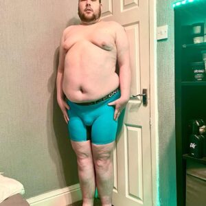 A tall bearded man wearing a pair of teal blue boxer briefs with a black waistband that has Step One written in bold blue writing on it stood against a white wooden door.