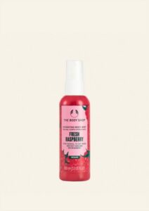 A tall cylindrical clear plastic Bottle full of some dark red liquid with a white plastic spray pump lid and a bright pink label that has The Body Shop Fresh Raspberry Hydrating Body Mist written in dark green writing and a picture of some dark red raspberries on it, on a white background.