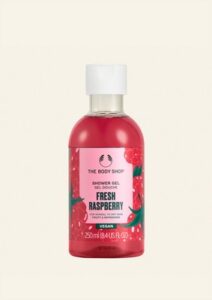 A tall cylindrical clear plastic bottle full of some dark red liquid with a white plastic lid and a bright pink label that has The Body Shop Fresh Raspberry Shower Gel written in dark green writing and a picture of some dark red raspberries on it, on a white background.
