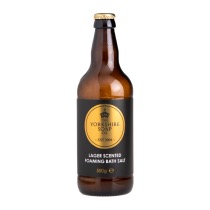 A tall cylindrical dark amber glass beer bottle with a metallic gold lid and a black label that has The Yorkshire Soap Company Lager Foaming Bath Salts written in bold white writing on it, on a white background.