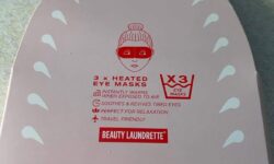 A large triangular bright pink cardboard box that has beauty Laundrette heated eye masks written in bold red writing and a picture of a dark red face wearing a bright Red eye mask on it, on a white background.
