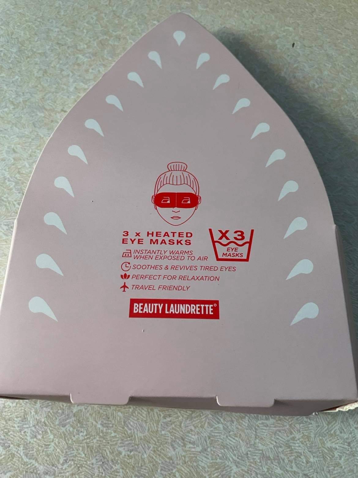 A large triangular bright pink cardboard box that has beauty Laundrette heated eye masks written in bold red writing and a picture of a dark red face wearing a bright Red eye mask on it, on a white background.