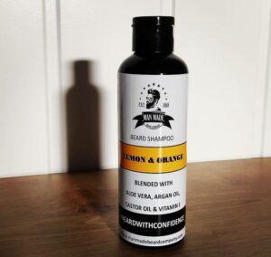 A tall cylindrical black Plastic Bottle with a black Plastic Lid and a white label that has Man Made Beard Company Lemon & Orange Beard Shampoo written in bold black writing and a picture of a man with a black beard on it, on a white background.
