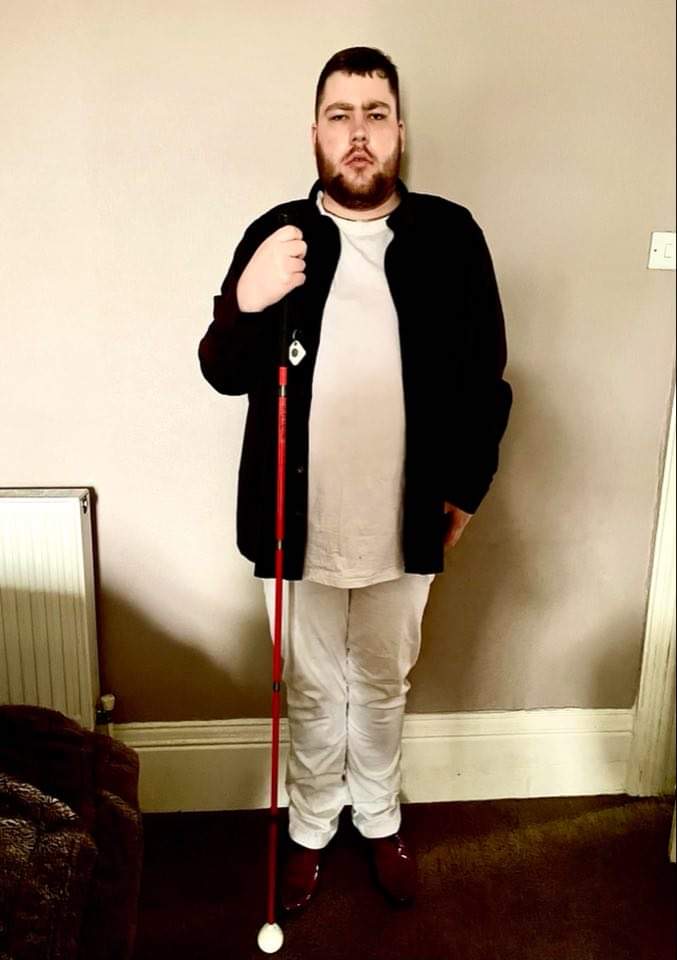 Outfit Of The Day | A Day Out In Liverpool A tall bearded man wearing a plum long sleeve oxford shirt, a bright white crew neck t-shirt, a pair of white chinos and a pair of shiny red shoes holding a bright red cane stood against a light grey wall.