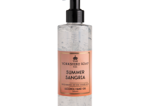 A tall cylindrical clear plastic bottle full of some translucent pink liquid with a black plastic pump lid and a bright pink label that has The Yorkshire Soap Company Summer Sangria Alcohol Hand Gel written in bold black writing on it, on a white background.