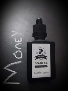 A short rectangular brown glass Bottle with a black rubber pipette lid and a white label that has Man Made Beard Company The Money Man Beard Oil written in bold black writing and a picture of a man with a black beard on it, on a white background.