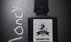 A short rectangular brown glass Bottle with a black rubber pipette lid and a white label that has Man Made Beard Company The Money Man Beard Oil written in bold black writing and a picture of a man with a black beard on it, on a white background.
