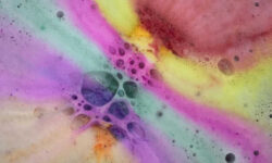 A large pile of fluffy rainbow coloured bubbles inside a bath full of water, on a white background.
