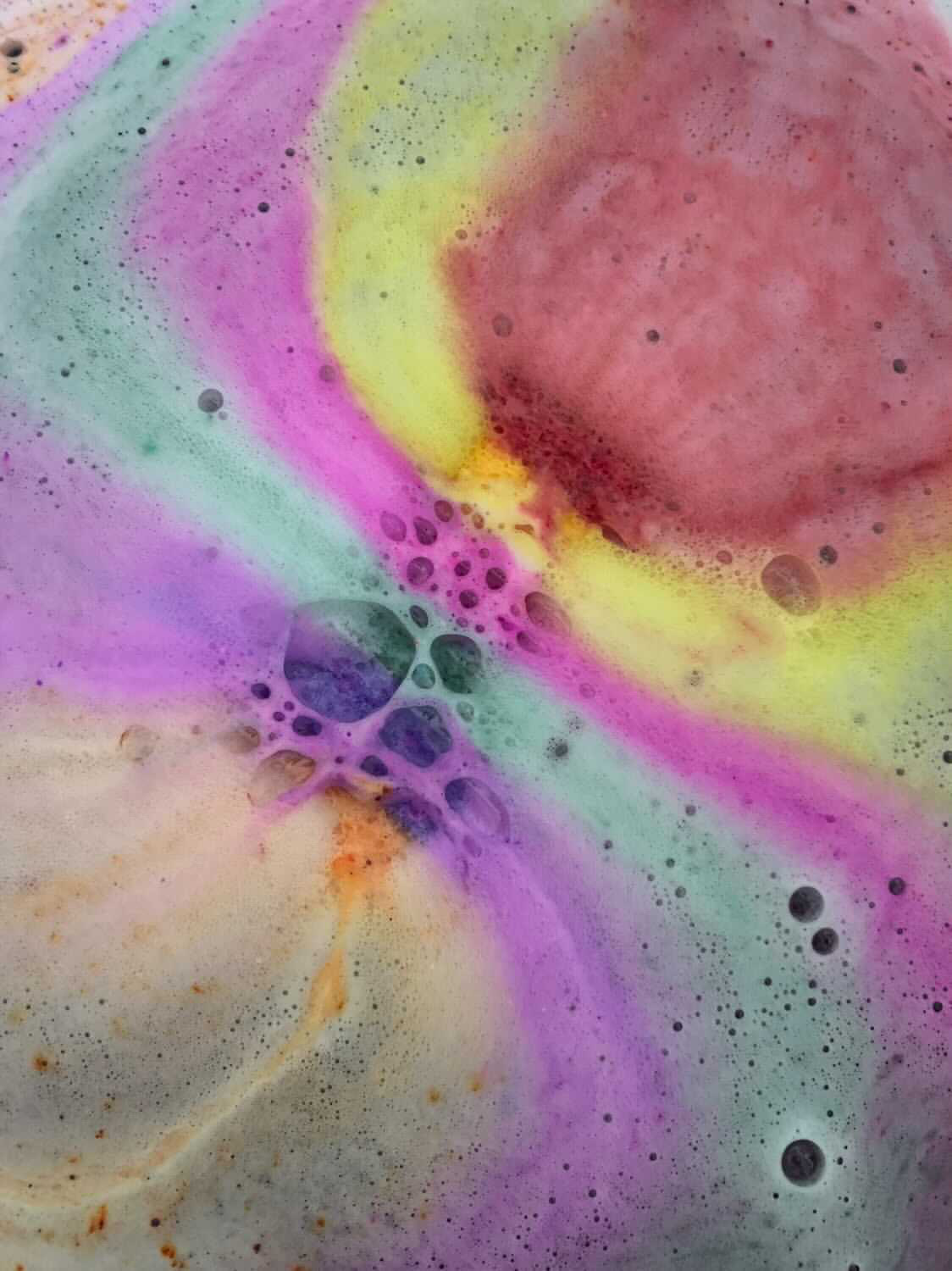 A large pile of fluffy rainbow coloured bubbles inside a bath full of water, on a white background.