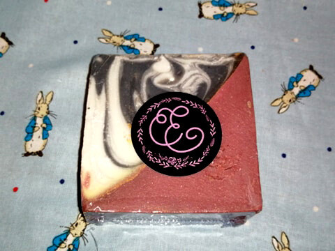 A large rectangular block of pale pink soap that has a black and white tornado design on it next to a rectangular black sticker that has Elpida Soaps written in bright pink writing on it, on a white background.