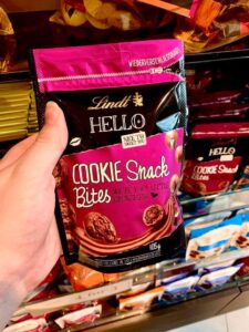 A tall rectangular gradient pink and brown plastic Pouch that has Lindt written in bold gold writing, Hello Cookie Snack Bites written in bold white writing, and a picture of some small spherical brown chocolate balls on it, on a white background.