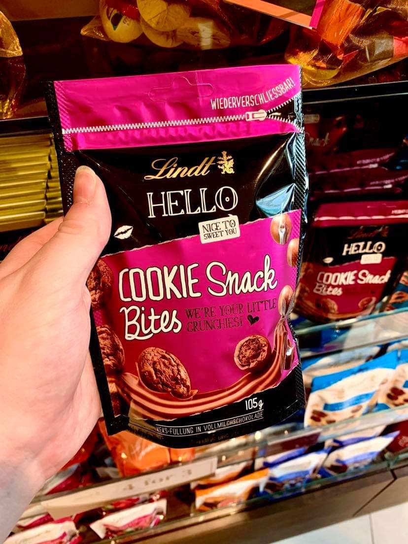 Say Hello To The Perfect Snack | Lindt Hello Snack Bites A tall rectangular gradient pink and brown plastic Pouch that has Lindt written in bold gold writing, Hello Cookie Snack Bites written in bold white writing, and a picture of some small spherical brown chocolate balls on it, on a white background.